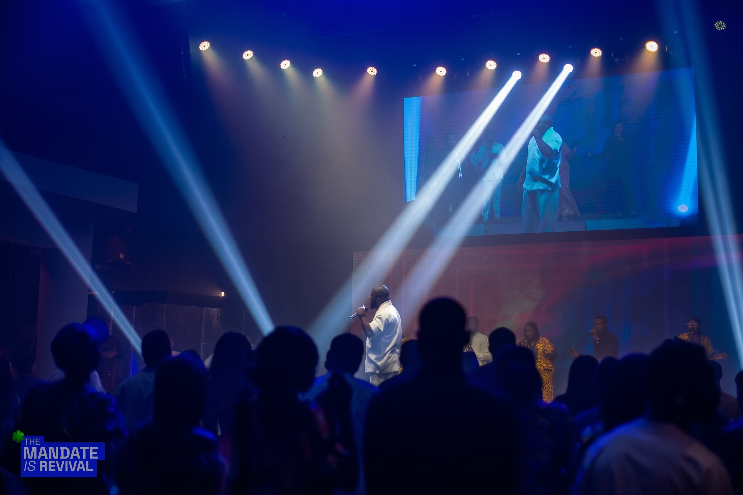 Samuel Folabi on a blue-lit stage, mirrored on the large screen above the crowd