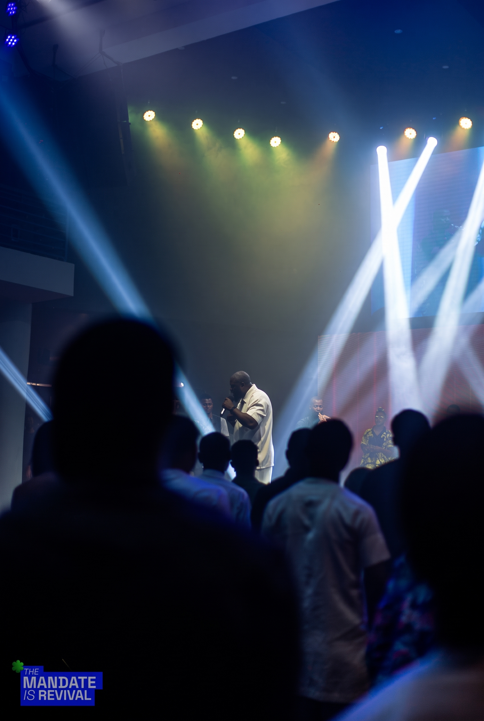 Samuel Folabi in prayer on stage, seen past the silhouettes of worshippers